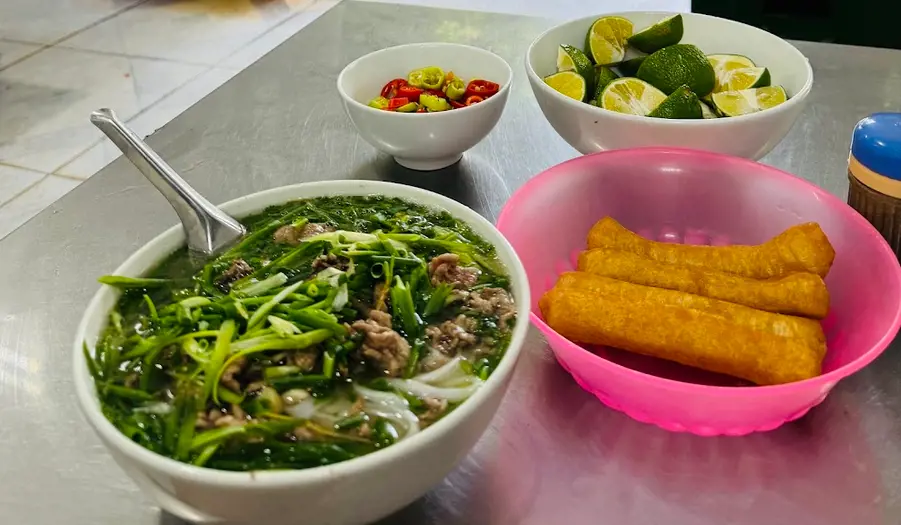 Bát phở bò Phở Thìn Hà Nội đầy hành lá và thịt bò, bên cạnh là quẩy, chanh và ớt thái lát trên bàn kim loại. – Cách thưởng thức trọn vẹn một tô phở